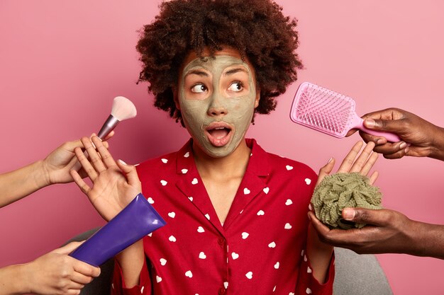 surprised-beautiful-woman-wears-red-night-clothes-applies-clay-mask-face-raises-palm-towards-various-beauty-products_273609-38708